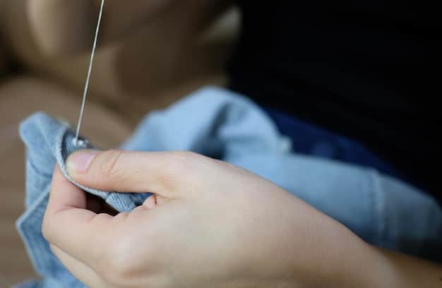 A close-up of hands mending a torn fabric with a needle and thread, showcasing the act of repair and prolonging garment life.